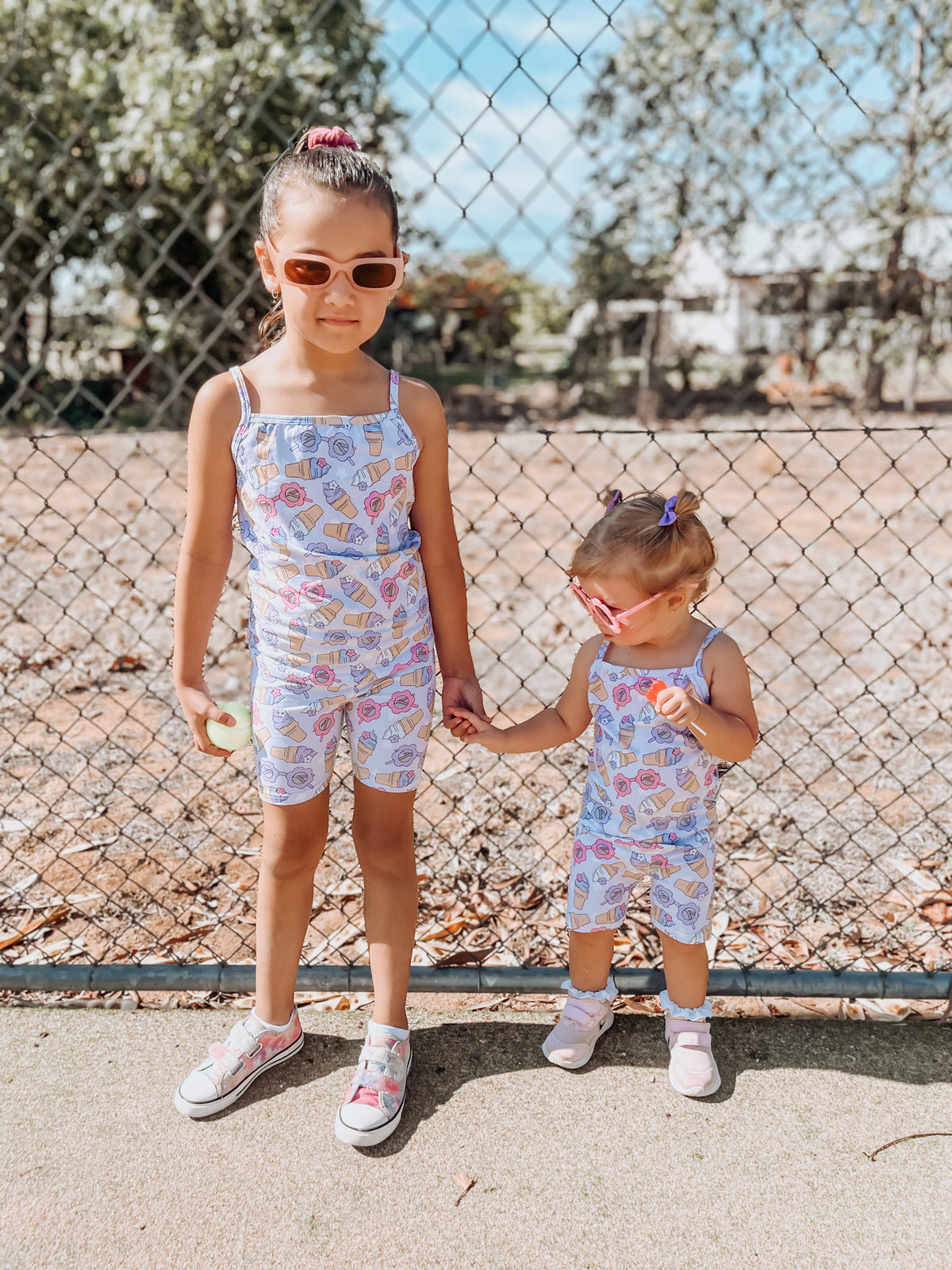 Two young girls standing next to a fence, wearing matching floral bike pants with sunglasses and shoes visible.
